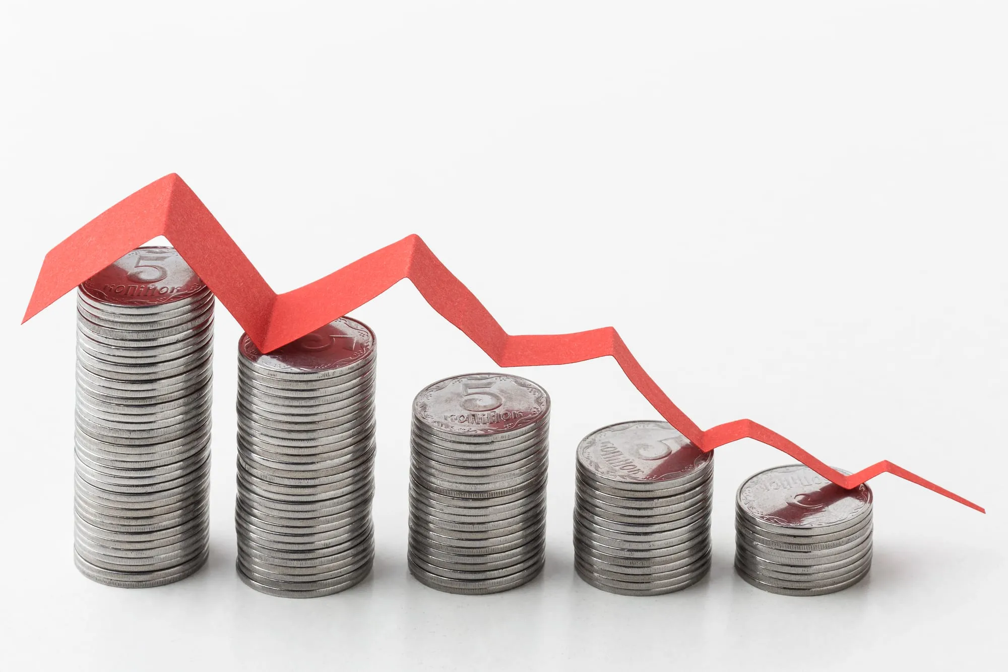 Five descending coin stacks with a red paper arrow trending downward across them, illustrating declining value.