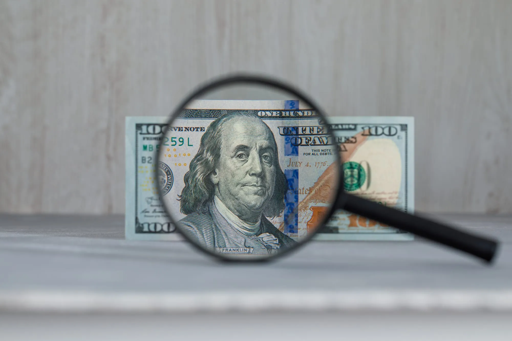 A magnifying glass held over a $100 bill, enlarging Benjamin Franklin's portrait, against a wood-grain background.