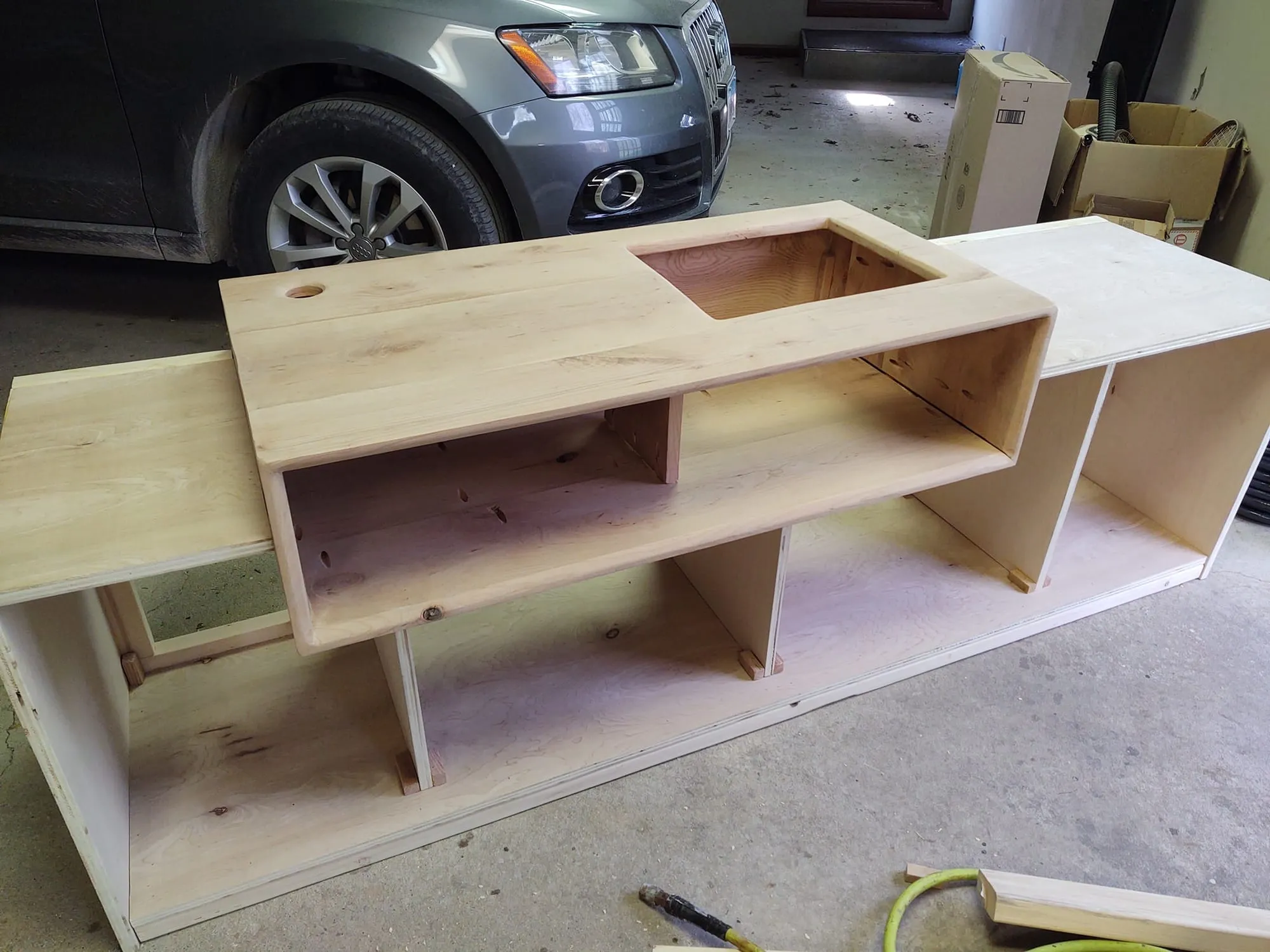 Unstained turntable stand under construction in a garage, with a car parked behind it and a shelf compartment with a rectangular cutout.