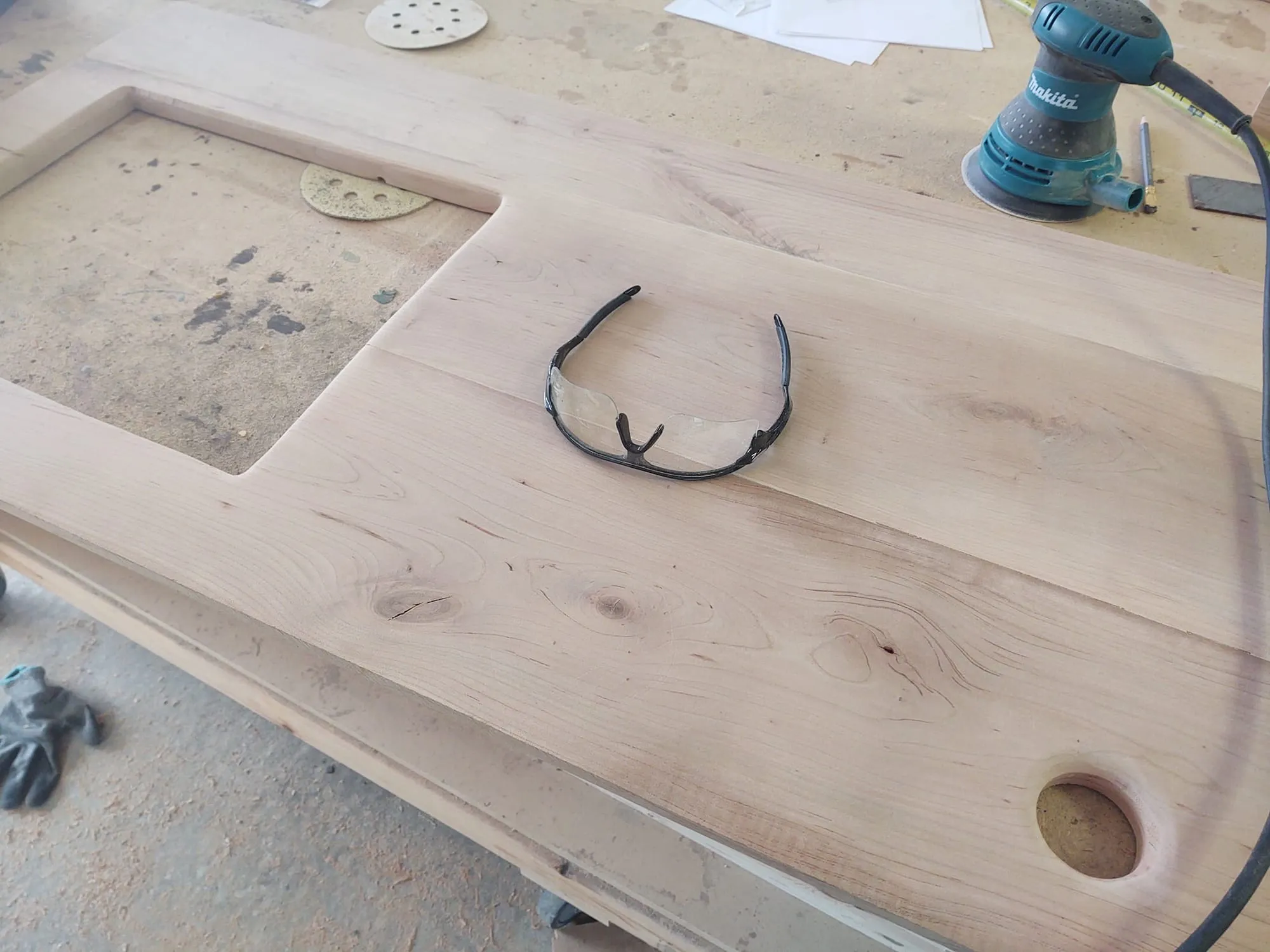 Unfinished turntable stand top made of light wood, with safety glasses resting on it beside a Makita orbital sander on a garage workbench.