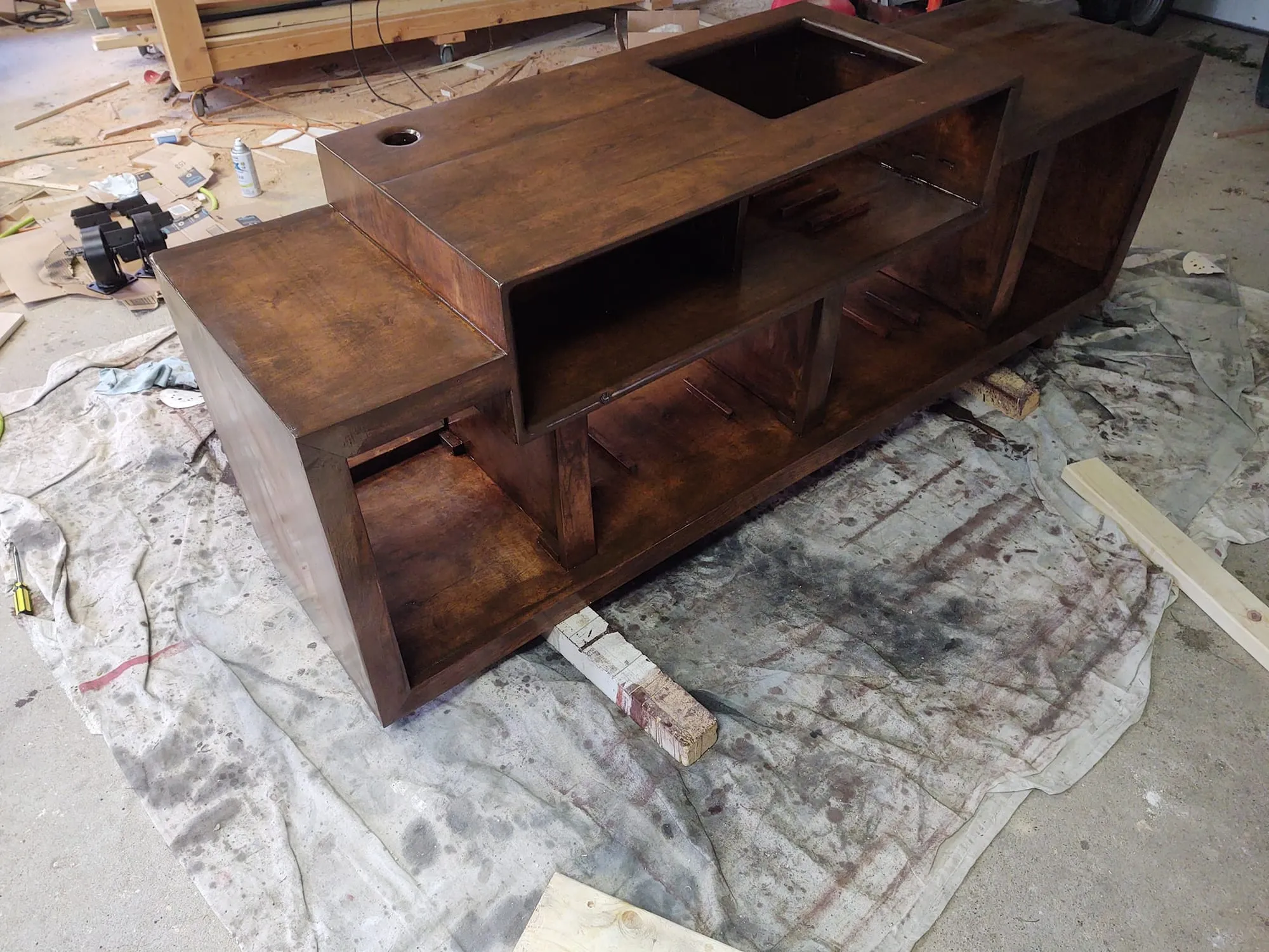 Dark-stained turntable stand assembled in a garage workshop, sitting on a drop cloth, showing open shelving and a rectangular cutout.