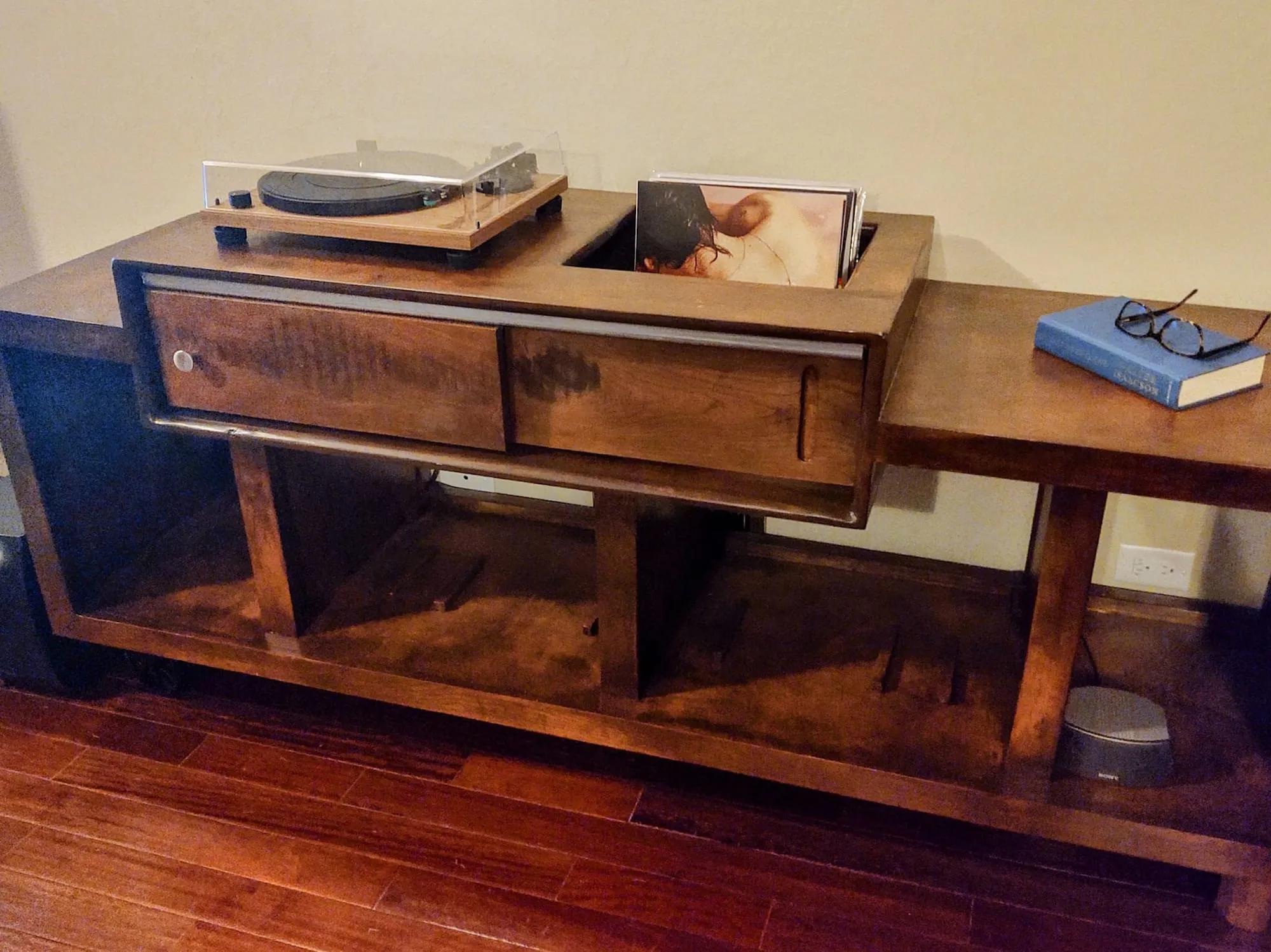 Finished dark wood turntable stand in a living room, with a turntable and vinyl records on top and a record-storage cutout below.